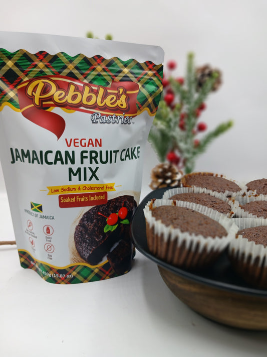 A bag of Pebble's Pastries Vegan Jamaican Fruit Cake Mix stands next to a bowl of fruit cake cupcakes on a white surface with a blurred holiday background.