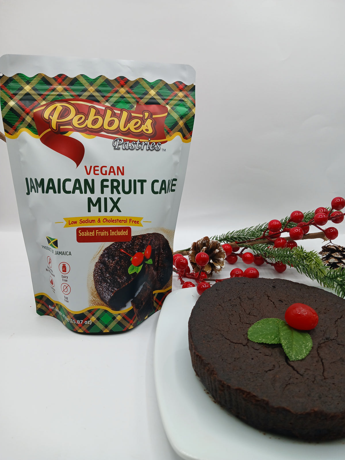 A pouch of Pebble's Pastries Vegan Jamaican Fruit Cake Mix stands next to a round fruit cake on a white plate, decorated with a green leaf and a red berry, with festive pine branches and red berries in the background.